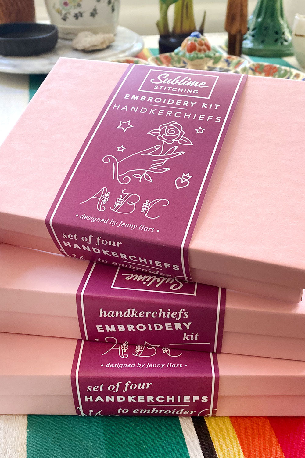A stack of pink boxes with plum-colored belly bands that read "Sublime Stitching Embroidery Kit: Handkerchiefs"
